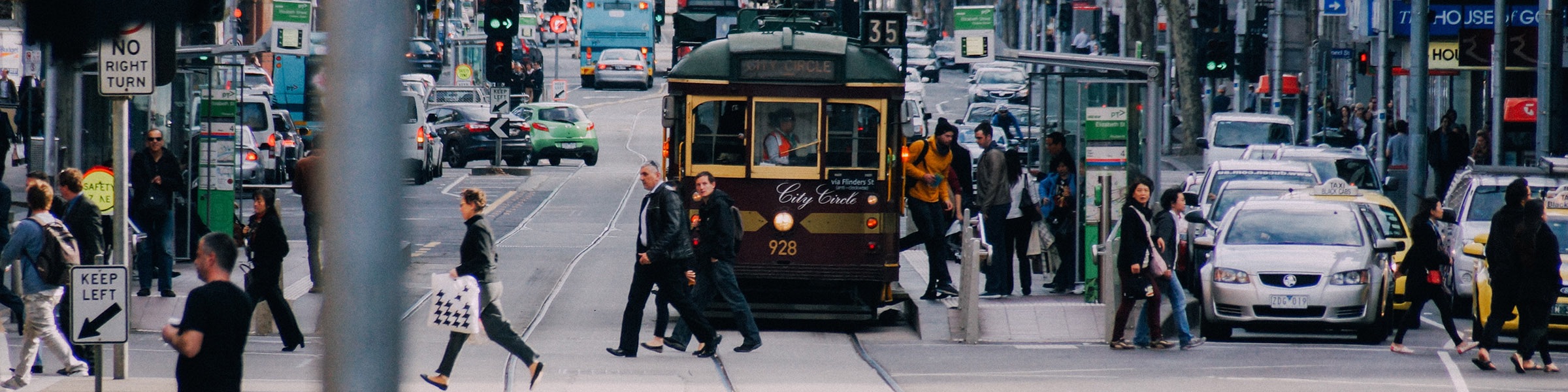 Trams A busy urban street scene features pedestrians crossing in front of a vintage tram while traffic flows in the background. People are seen walking with bags, waiting at a bus stop, and navigating the crosswalks amidst a variety of vehicles, including cars and taxis.